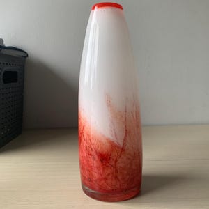 Peut inclure: Un vase cylindrique en verre haut, présentant une partie supérieure blanche lisse qui se fond dans une base rouge vif. La partie inférieure affiche un motif marbré veiné en rouge profond, tandis que le bord est orné d'une bande rouge.