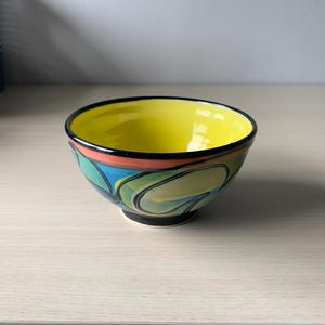 May include: A ceramic bowl featuring a bright yellow interior and a black rim. The exterior displays a colourful abstract pattern with curved shapes in shades of teal, light green, and orange, all outlined in bold black lines against a dark base.