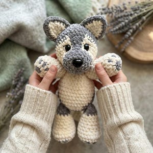 May include: A handmade crochet plush toy shaped like a wolf or husky puppy. The stuffed animal features a soft, textured yarn construction with a color palette of cream and grey. It has two black plastic eyes, a black nose, and pointed ears.