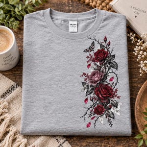 Pode incluir: Uma t-shirt cinzenta mesclada de gola redonda com um gráfico floral vertical no lado direito. O design inclui rosas vermelho-escuras, caules espinhosos, folhas verde-escuras e uma pequena traça. Etiqueta da marca Gildan no interior da gola.