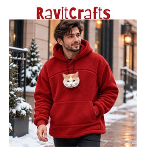 Op de afbeelding: Een rode fleece hoodie met een grote voorzak en een afbeelding van een rood-witte kattenkop in het midden op de borst. De hoodie heeft een capuchon met zwarte trekkoorden en lange mouwen met geribbelde boorden. De tekst RavitCrafts staat bovenaan.
