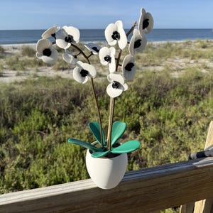 May include: A decorative artificial orchid plant featuring white petals with black centers, supported by thin brown stems. The plant has several smooth, vibrant green leaves at the base and is potted in a simple, matte white rounded container.