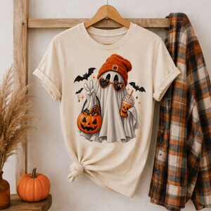 Op de afbeelding: Een crèmekleurig T-shirt met korte mouwen en een afbeelding van een geest met een oranje muts en hartvormige zonnebril. De geest houdt een pompoenvormige snoepemmer en een drankje vast, omringd door kleine vleermuizen en steraccenten.