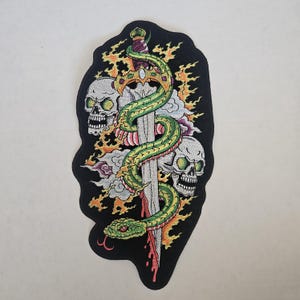 May include: An embroidered patch on a black background featuring a silver sword wrapped in a green and yellow snake. Two white skulls are positioned on either side of the blade, surrounded by orange flames and light purple cloud-like shapes.