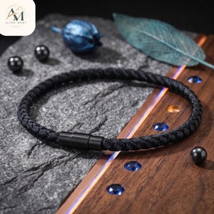 May include: A black braided cord bracelet featuring a cylindrical matte black metal magnetic clasp. The bracelet is circular in shape and displays a consistent woven texture throughout the entire length of the cord, providing a uniform appearance.