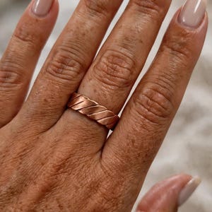 Op de afbeelding: Een koperen ring met een doorlopend diagonaal gedraaid of gevlochten patroon rondom de gehele band. Het metaal heeft een warme, roodbruine metallic afwerking met een gepolijst oppervlak dat het licht reflecteert langs de schuine randen.