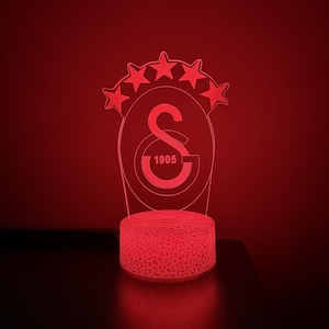 Pode incluir: Uma lâmpada LED vermelha com um painel de acrílico transparente exibindo o logótipo do clube desportivo Galatasaray, incluindo a letra S, o ano 1905 e cinco estrelas no topo. A base circular tem um padrão texturado e rachado iluminado em vermelho.