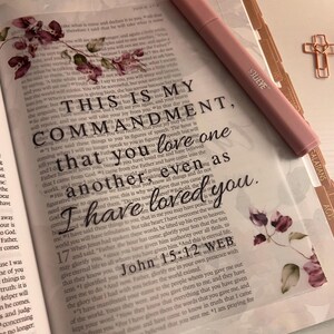May include: An open book with a transparent overlay featuring the text: THIS IS MY COMMANDMENT, that you love one another, even as I have loved you. John 15:12 WEB. A pink highlighter and a gold cross-shaped paperclip rest on the page.