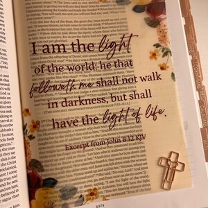 May include: An open Bible page featuring floral illustrations in the corners and the text: I am the light of the world: he that followeth me shall not walk in darkness, but shall have the light of life. Excerpt from John 8:12 KJV. A gold cross paperclip is attached.