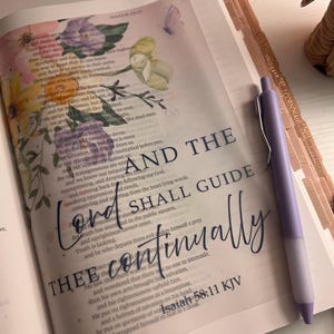 May include: An open Bible page featuring floral illustrations and a butterfly, with the text overlay "AND THE Lord SHALL GUIDE THEE continually" and "Isaiah 58:11 KJV" printed in black. A light purple retractable pen rests on the right side of the page.