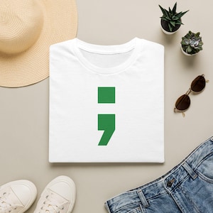 Könnte beinhalten: Ein weißes T-Shirt mit einem großen, grünen Semikolon-Grafikdruck mittig auf der Brust. Das Shirt ist zusammen mit einem Strohhut, zwei kleinen Sukkulenten in Töpfen, einer braunen Sonnenbrille, weißen Turnschuhen und einer blauen Jeans abgebildet.