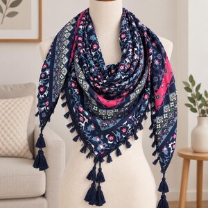 Pode incluir: Um lenço triangular com uma base azul-escura e um padrão floral e geométrico vibrante em tons de rosa, branco e azul-claro. As bordas são finalizadas com uma série de pequenas borlas azul-escuras ao longo da extremidade inferior.