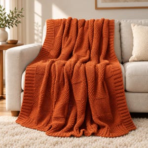 May include: A burnt orange knitted throw blanket featuring a textured checkerboard pattern with alternating smooth and bumpy squares, finished with a wide ribbed border along the edges, draped over the seat of a light-colored sofa.