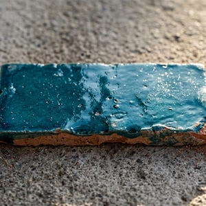 Peut inclure: Trois carreaux de céramique rectangulaires avec une glaçure bleu sarcelle profonde et brillante et des bords en terre cuite visibles. Les carreaux présentent une texture de surface inégale et artisanale avec des variations subtiles.