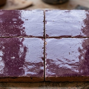 May include: Four square ceramic tiles arranged in a grid, each featuring a deep, glossy purple glaze with a visible crackle finish. The tiles have a thick, earthy terracotta base visible along the edges, showcasing a handcrafted, rustic texture.