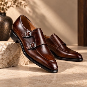 Pode incluir: Um par de sapatos de couro castanho escuro com fivela dupla, acabamento polido, fivelas de metal prateado e sola preta. Os sapatos apresentam um design de bico fino e uma parte superior de couro liso polido com detalhes de costura.