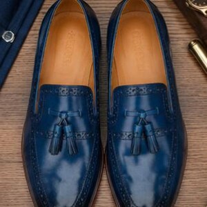 May include: A pair of deep blue leather loafers featuring a tassel detail on the vamp, decorative brogue perforations along the edges, and a tan leather interior lining. The shoes have a smooth, polished finish and a rounded toe silhouette.