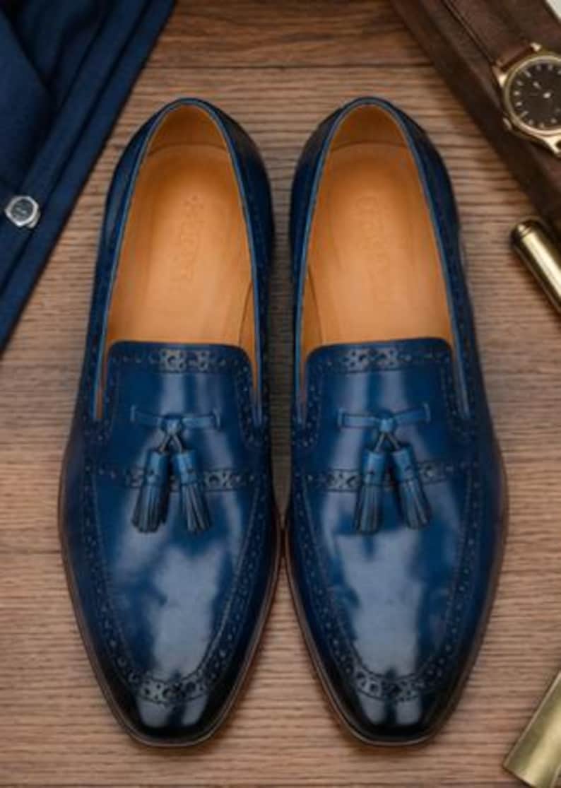 May include: A pair of deep blue leather loafers featuring a tassel detail on the vamp, decorative brogue perforations along the edges, and a tan leather interior lining. The shoes have a smooth, polished finish and a rounded toe silhouette.