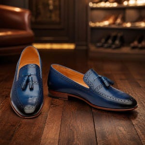 May include: A pair of deep blue leather tassel loafers featuring intricate brogue detailing along the edges, a smooth polished finish, and a stacked wooden heel. The shoes have a tan interior lining and a rounded toe design with a dark gradient.