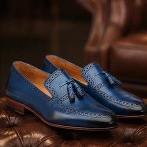 May include: A pair of blue leather loafers featuring a dark gradient finish at the toe, decorative tassels on the vamp, and intricate brogue detailing along the edges. The shoes have a tan interior lining and a stacked brown wooden heel.