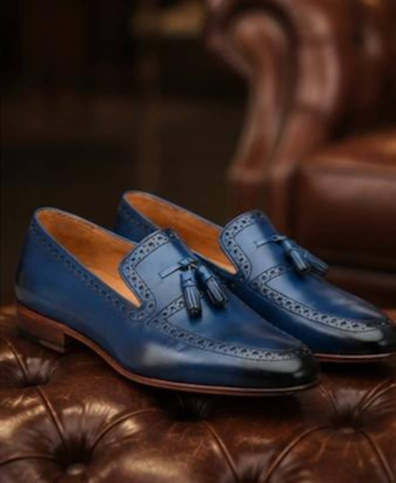 May include: A pair of blue leather loafers featuring a dark gradient finish at the toe, decorative tassels on the vamp, and intricate brogue detailing along the edges. The shoes have a tan interior lining and a stacked brown wooden heel.