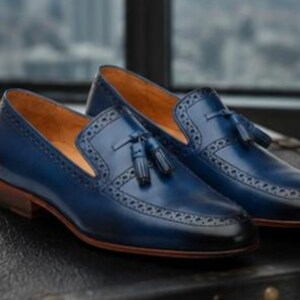 May include: A pair of navy blue leather tassel loafers featuring a burnished toe, intricate brogue detailing along the vamp, and a tan leather interior lining. The shoes have a low stacked heel and a smooth finish with a classic slip-on design.