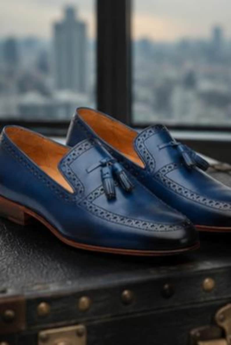 May include: A pair of navy blue leather tassel loafers featuring a burnished toe, intricate brogue detailing along the vamp, and a tan leather interior lining. The shoes have a low stacked heel and a smooth finish with a classic slip-on design.
