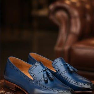 May include: A pair of blue leather tassel loafers featuring decorative brogue detailing along the edges and a smooth finish. The shoes have a low heel, a tan interior lining, and a dark gradient effect at the rounded toe and heel areas.