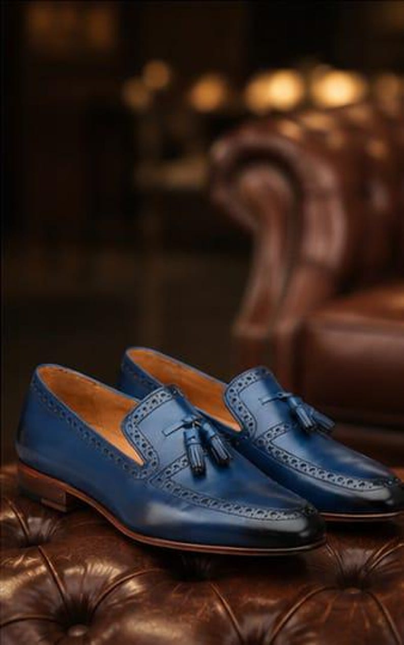 May include: A pair of blue leather tassel loafers featuring decorative brogue detailing along the edges and a smooth finish. The shoes have a low heel, a tan interior lining, and a dark gradient effect at the rounded toe and heel areas.