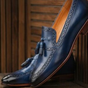 May include: A pair of navy blue leather loafers featuring a tassel detail on the vamp, intricate brogue perforations along the edges, a pointed toe, and a stacked wooden heel. The interior lining is a tan color, and the leather has a polished finish.