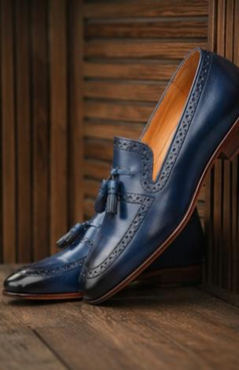 May include: A pair of navy blue leather loafers featuring a tassel detail on the vamp, intricate brogue perforations along the edges, a pointed toe, and a stacked wooden heel. The interior lining is a tan color, and the leather has a polished finish.