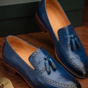 May include: A pair of blue leather tassel loafers featuring brogue detailing along the vamp and toe cap. The shoes have a tan interior lining, a dark brown stacked heel, and a gradient finish that transitions to black at the pointed toe.