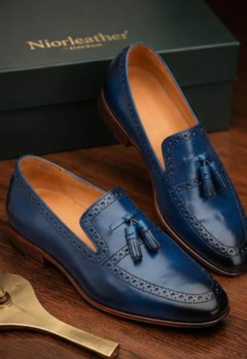 May include: A pair of blue leather tassel loafers featuring brogue detailing along the vamp and toe cap. The shoes have a tan interior lining, a dark brown stacked heel, and a gradient finish that transitions to black at the pointed toe.