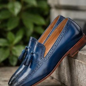 May include: A pair of deep blue leather loafers featuring decorative brogue detailing along the edges and a matching tassel accent on the vamp. The shoes have a smooth polished finish, a tan interior lining, and a stacked brown wooden heel.