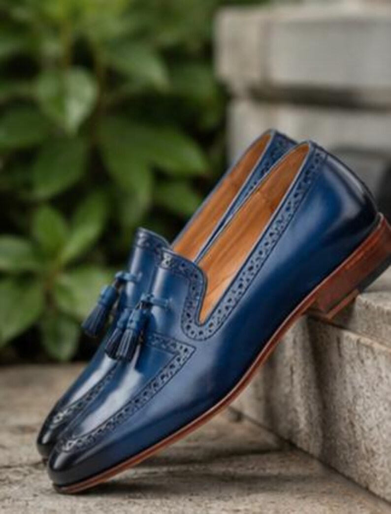 May include: A pair of deep blue leather loafers featuring decorative brogue detailing along the edges and a matching tassel accent on the vamp. The shoes have a smooth polished finish, a tan interior lining, and a stacked brown wooden heel.
