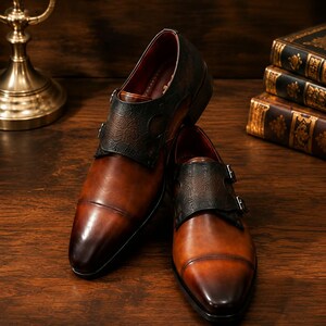Pode incluir: Um par de sapatos de couro castanho com fivelas, apresentando um painel de couro texturizado castanho escuro no peito do pé com duas fivelas prateadas. Os sapatos têm um acabamento polido, biqueira reforçada e forro interior vermelho escuro.