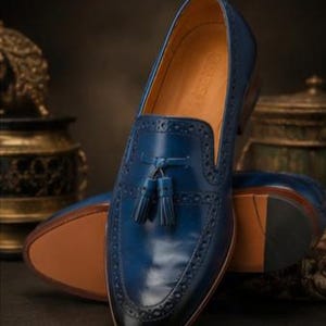 May include: A deep blue leather loafer featuring a decorative tassel on the vamp, intricate brogue detailing along the edges, a smooth tan interior lining, and a dark, polished toe cap, presented as a pair with one shoe standing upright.
