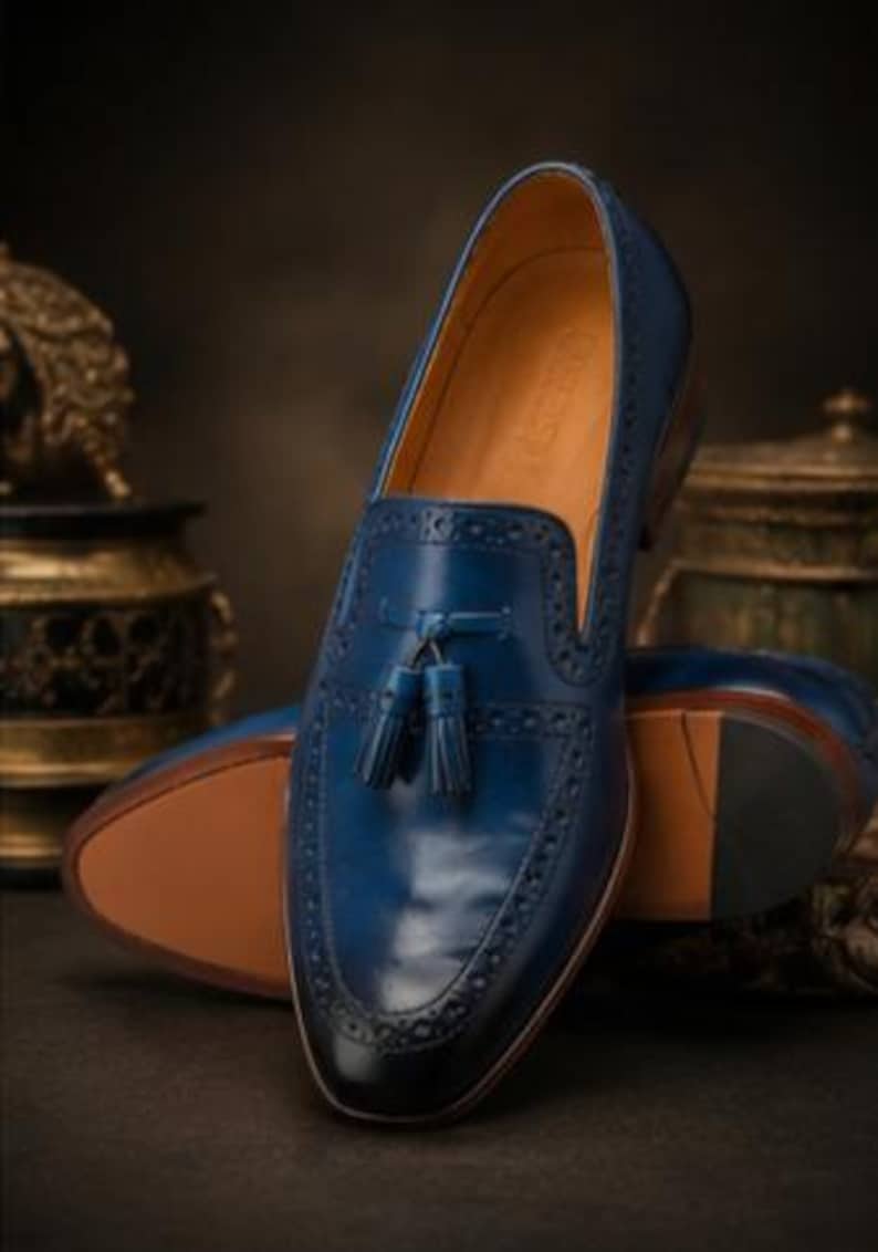 May include: A deep blue leather loafer featuring a decorative tassel on the vamp, intricate brogue detailing along the edges, a smooth tan interior lining, and a dark, polished toe cap, presented as a pair with one shoe standing upright.