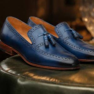 May include: A pair of navy blue leather loafers featuring a tassel detail on the vamp, intricate brogue perforations along the edges, a tan leather interior lining, and a stacked brown wooden heel with a matching brown leather sole.