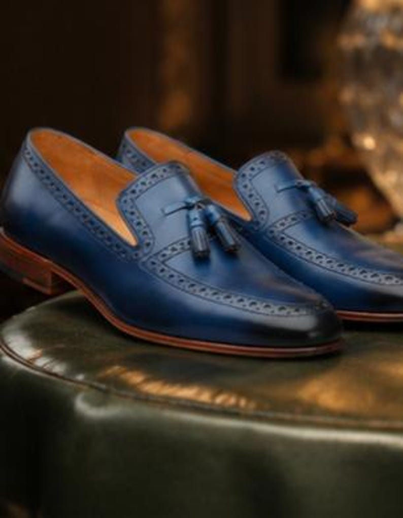 May include: A pair of navy blue leather loafers featuring a tassel detail on the vamp, intricate brogue perforations along the edges, a tan leather interior lining, and a stacked brown wooden heel with a matching brown leather sole.