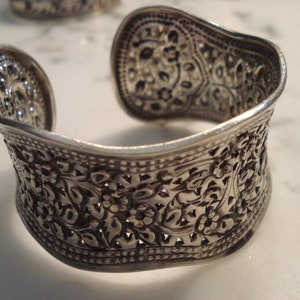 May include: A silver bracelet with an intricate floral design. The bracelet is open and has a wide, flat band.