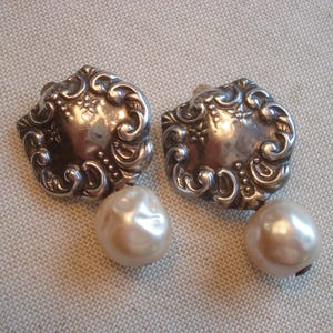 May include: A pair of silver-toned, ornate clip-on earrings. Each earring features a detailed, scrolled design with a central floral element and a large, pearlescent, round faux pearl drop. The earrings are displayed on a neutral, textured surface.