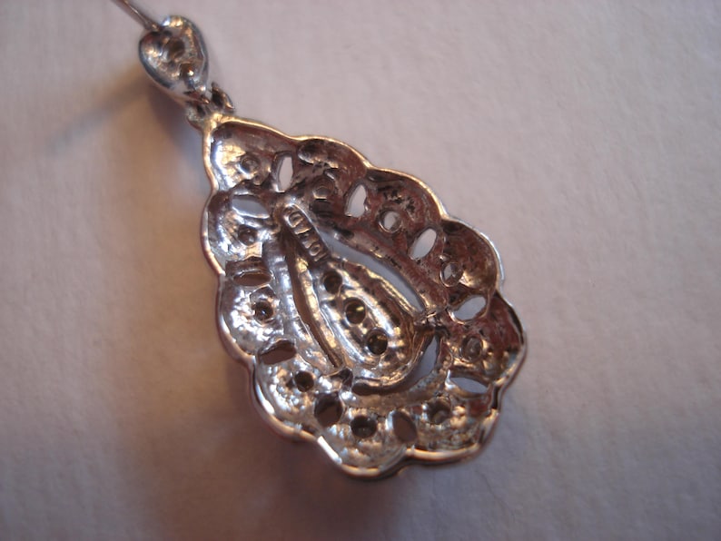 May include: Silver teardrop pendant with intricate filigree design and a small, clear stone in the center. The back of the pendant has a small, engraved mark.