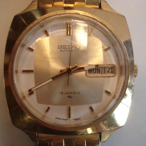 May include: A gold-toned wristwatch with a square face. The watch face is white with black markings and a date window that reads "MON 14". The watch face also has the text "SEIKO AUTOMATIC 19 JEWELS".