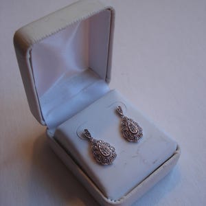May include: A pair of silver teardrop earrings with clear stones set in a delicate filigree design. The earrings are displayed in a white jewelry box.