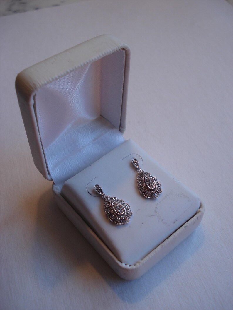 May include: A pair of silver teardrop earrings with clear stones set in a delicate filigree design. The earrings are displayed in a white jewelry box.