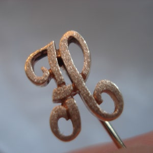 May include: Gold-toned metal pin with a stylized letter 'R' design. The pin has a textured surface and a thin, straight pin back.