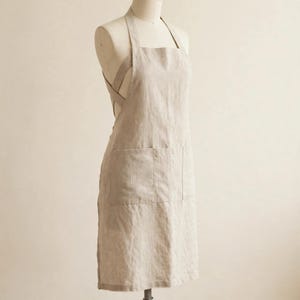 May include: A beige apron featuring a cross-back design with wide shoulder straps and two large front pockets. The apron is displayed on a mannequin, showing a simple, functional construction with a textured fabric finish.