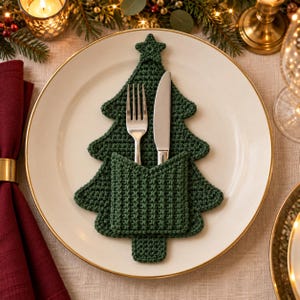 Peut inclure: Un porte-couverts en crochet vert foncé en forme de sapin de Noël avec une étoile au sommet. L'accessoire présente une poche sur le devant contenant une fourchette et un couteau en métal argenté, le tout centré sur une assiette blanche à bord doré.