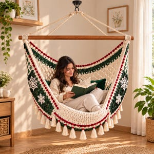 Puede incluir: Una silla colgante con una red de macramé color crema con detalles en verde oscuro, motivos de copos de nieve blancos y borde rojo. La silla está suspendida de una barra de madera horizontal y tiene borlas decorativas color crema.